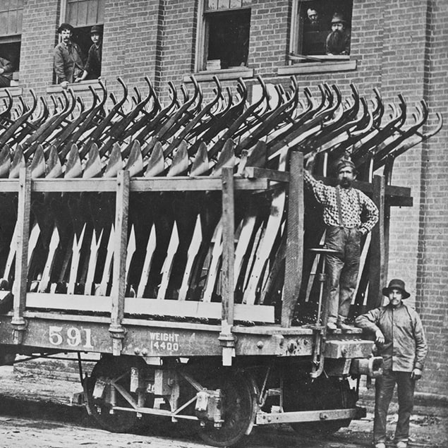 Zdjęcie z roku 1882 przedstawiające trzech mężczyzn stojących obok wagonu Deere & Co załadowanego stalowymi pługami gotowymi do transportu oraz pracowników fabryki patrzących przez okno znajdującego się na drugim planie budynku.