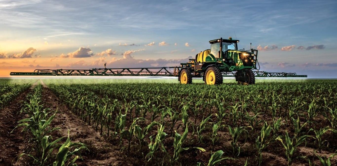 Opryskiwacz firmy John Deere pracujący na polu kukurydzy, w tle zachodzące słońce