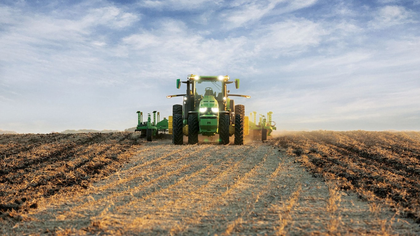 Samojezdny ciągnik John Deere ciągnący sprzęt do uprawy gleby przez otwarte pole.