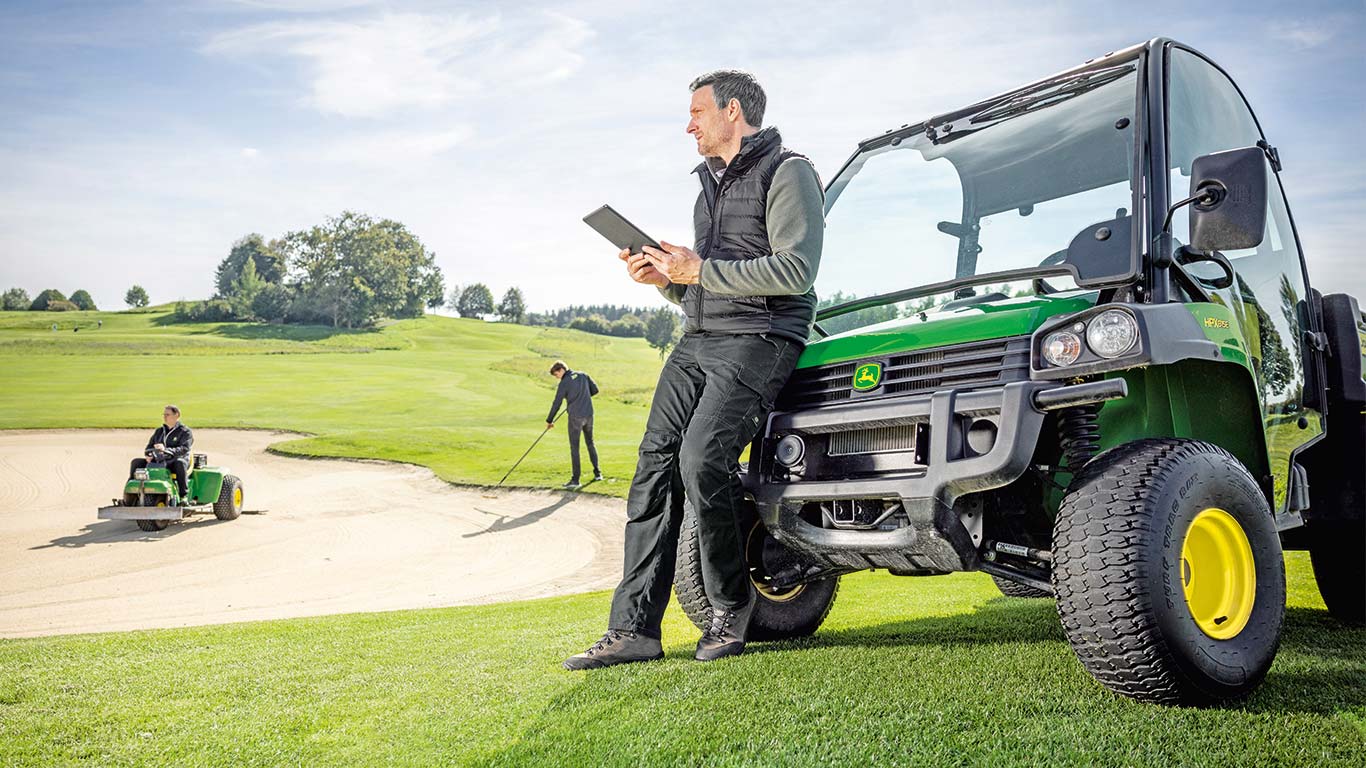 Główny greenkeeper pól golfowych sprawdza dane na tablecie, a 2 inni konserwatorzy pracują na polu golfowym w tle.