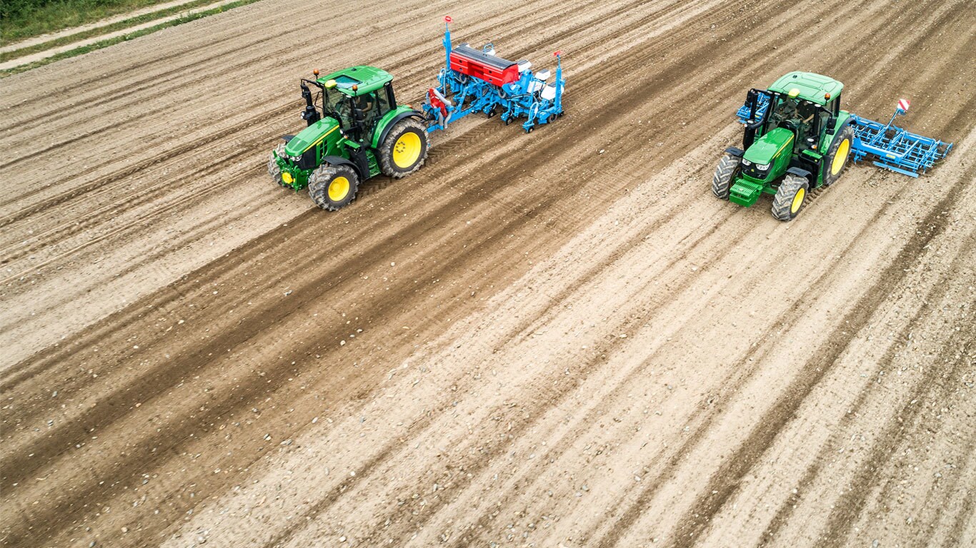 Dwa ciągniki John Deere 6M na gołej ziemi na polu. Jeden ciągnie siewnik Monsomen. Drugi ciągnie agregat uprawowy Lemken.