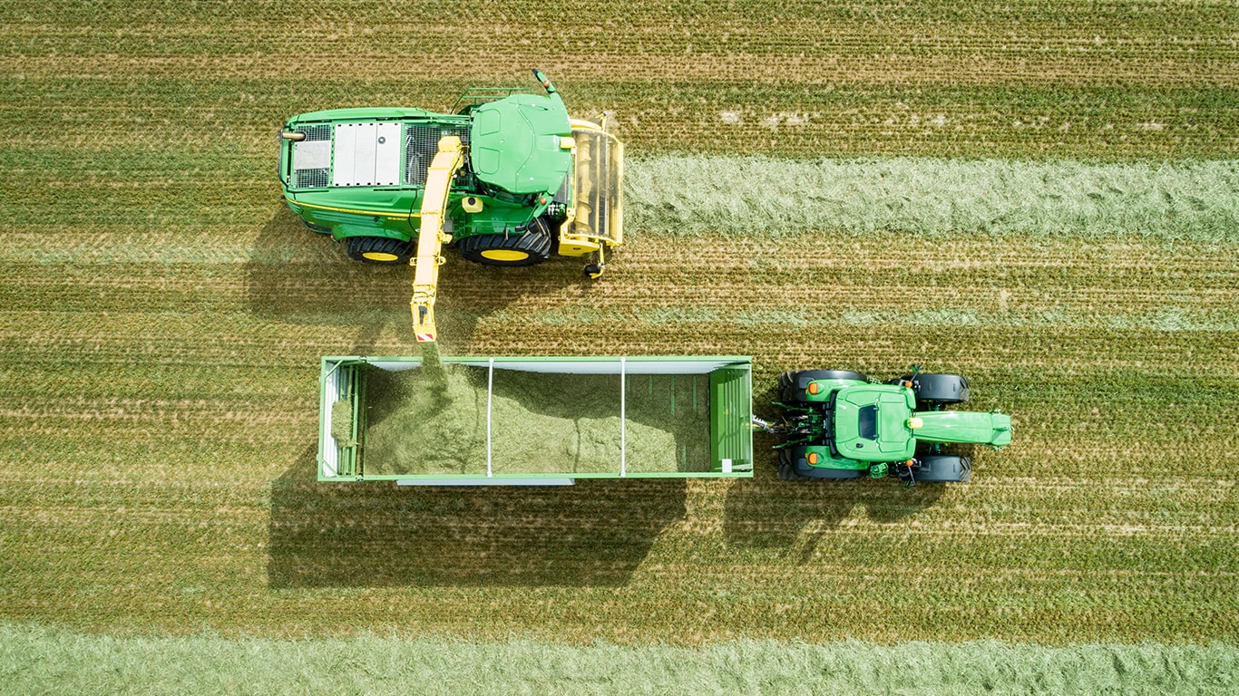 Zdjęcie z&nbsp;powietrza samojezdnej sieczkarni polowej John Deere serii&nbsp;8000 z&nbsp;podbieraczem trawy, ładującej kiszonkę na przyczepę ciągniętą przez ciągnik John Deere